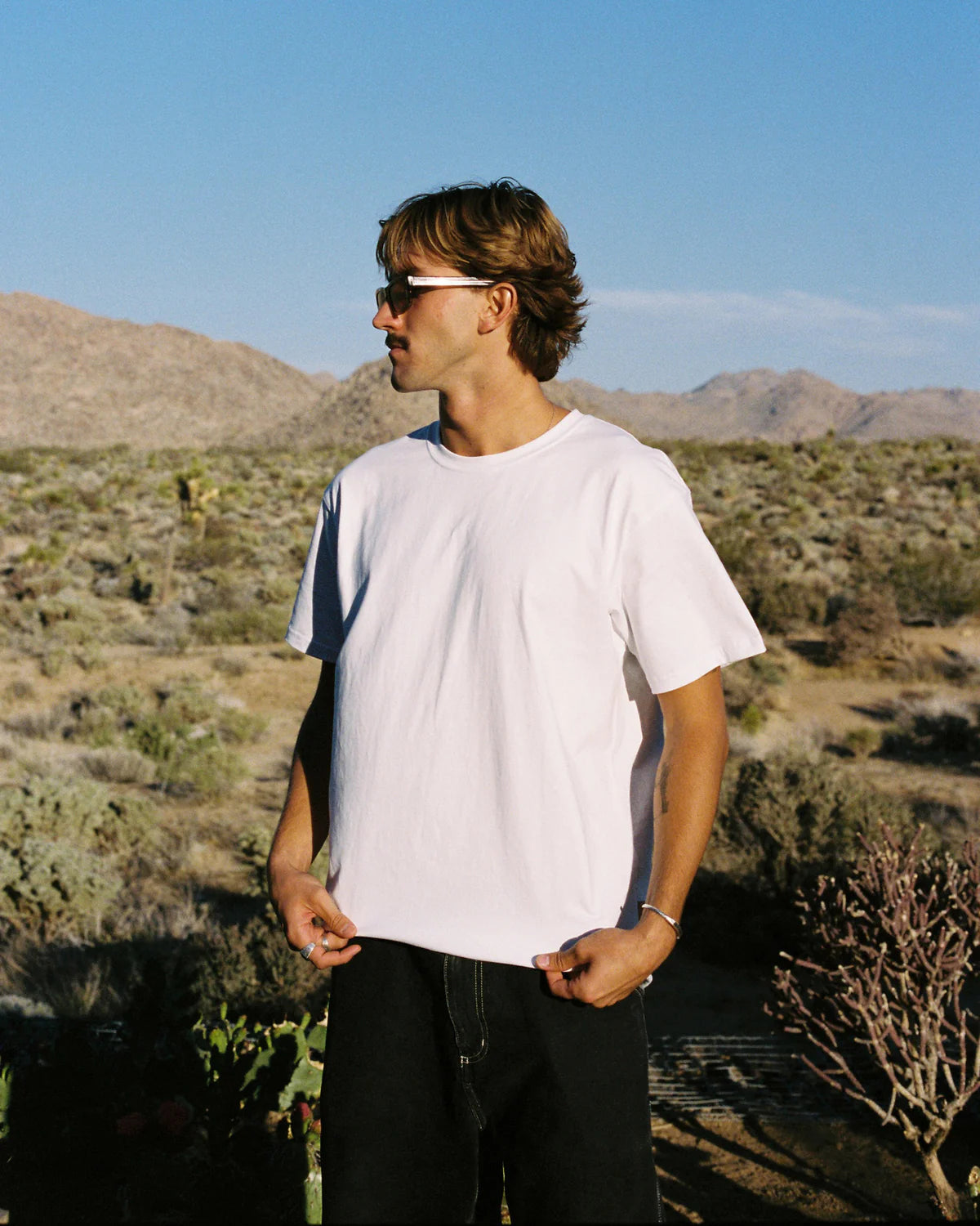 Man standing in a desert landscape wearing sunglasses and a white t-shirt.