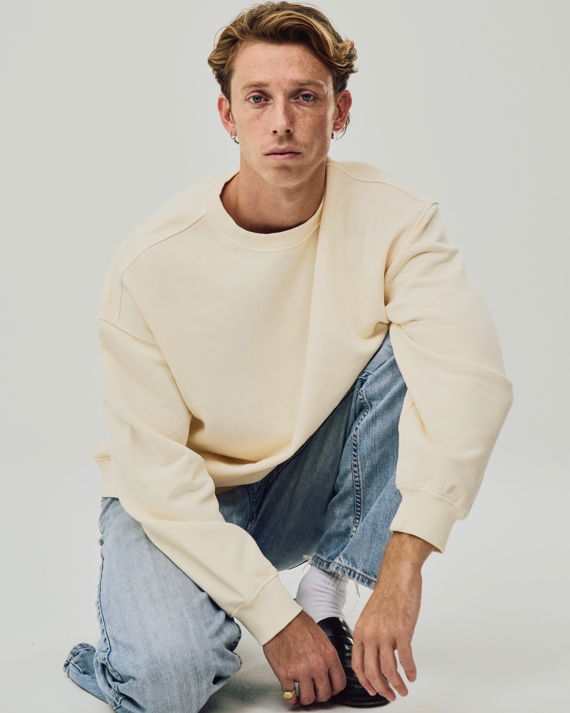 Man wearing a beige sweater and light blue jeans on a plain background