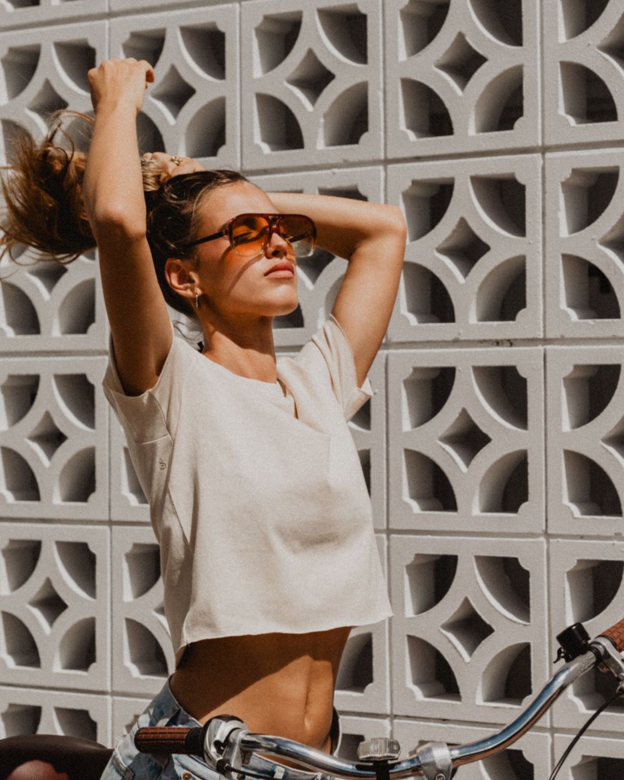 Woman with sunglasses standing next to a bicycle against a patterned wall.