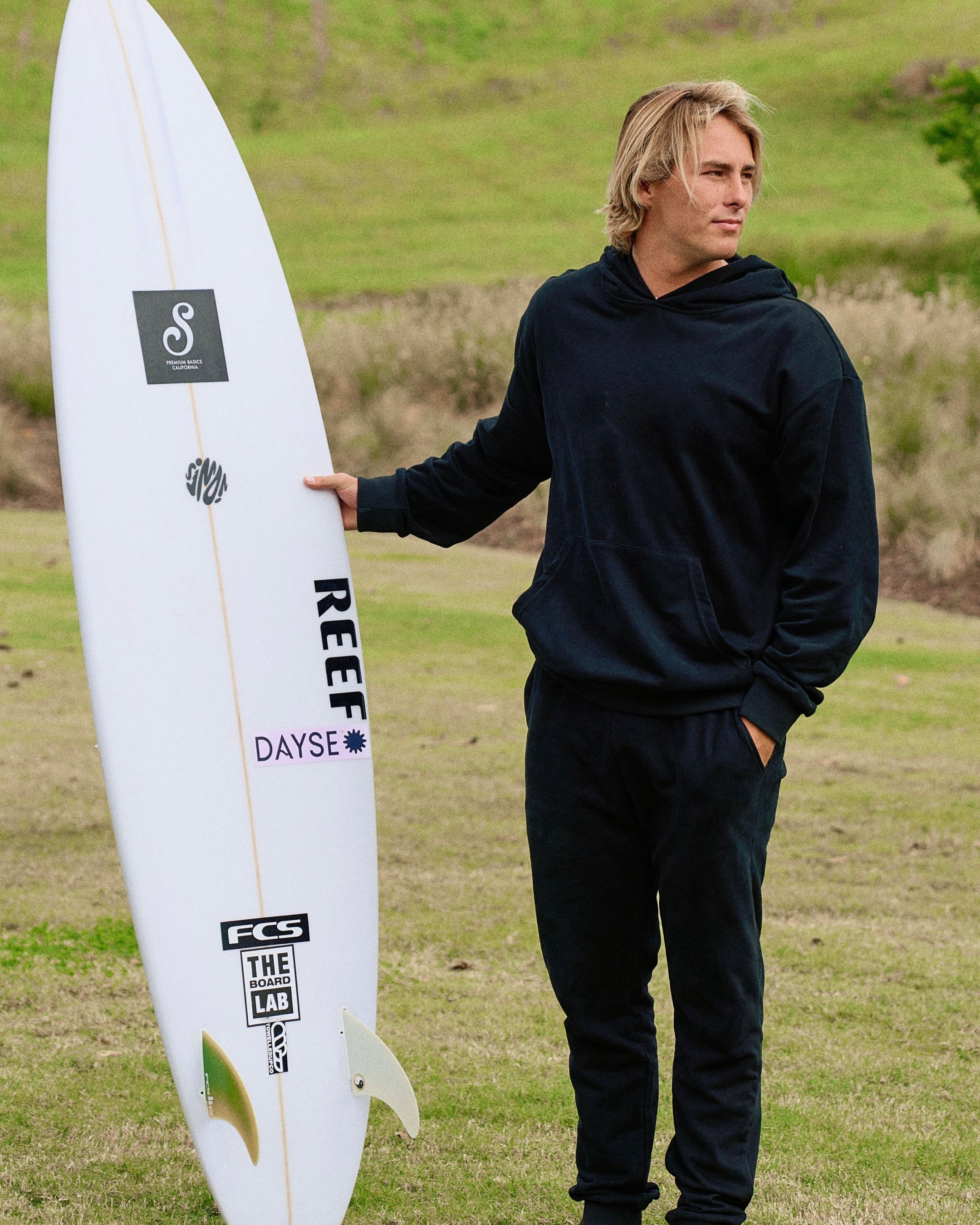 Man holding a white surfboard with branding in a grassy outdoor setting