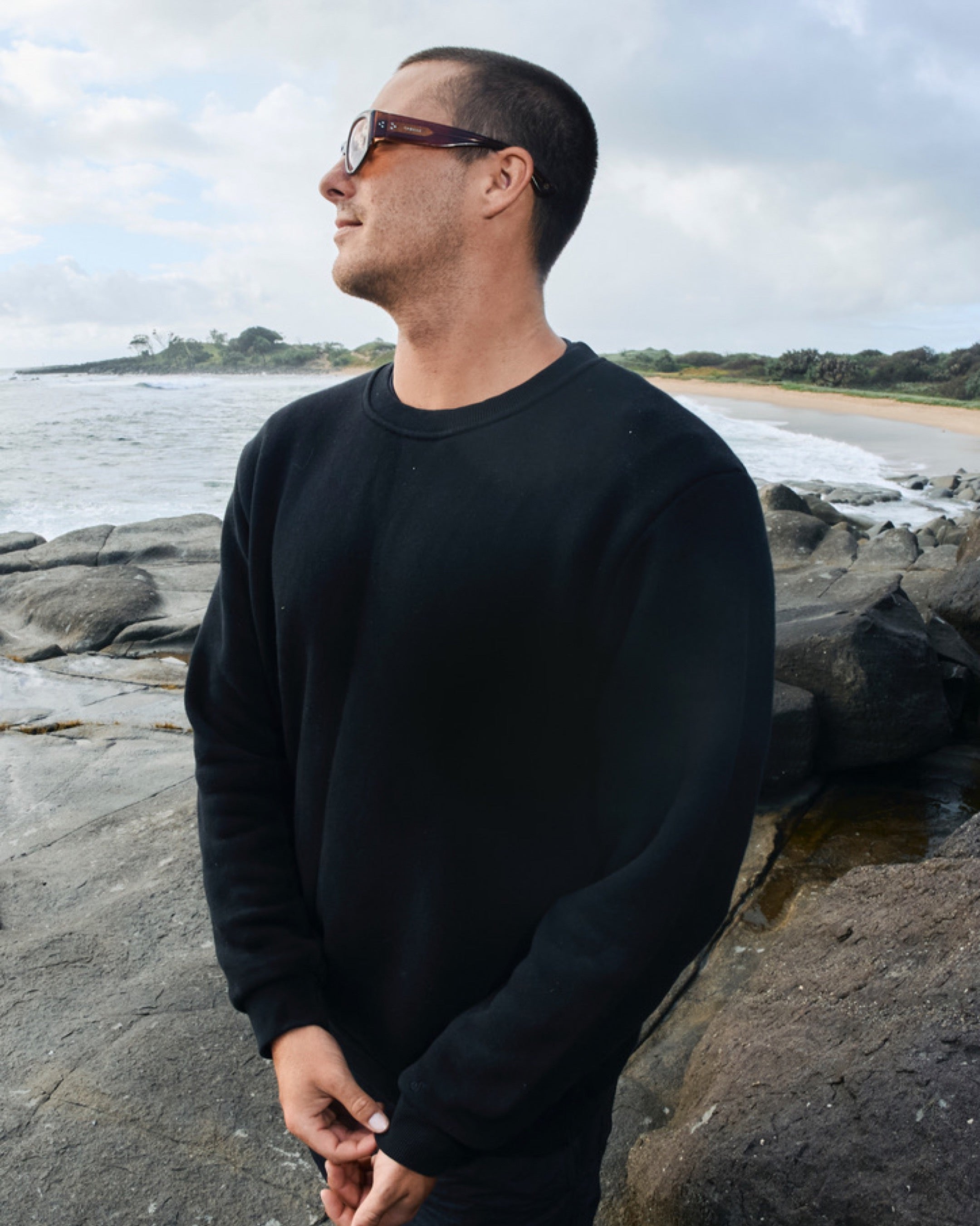 Man wearing a black sweatshirt standing on a rocky beach with ocean and sky in the background