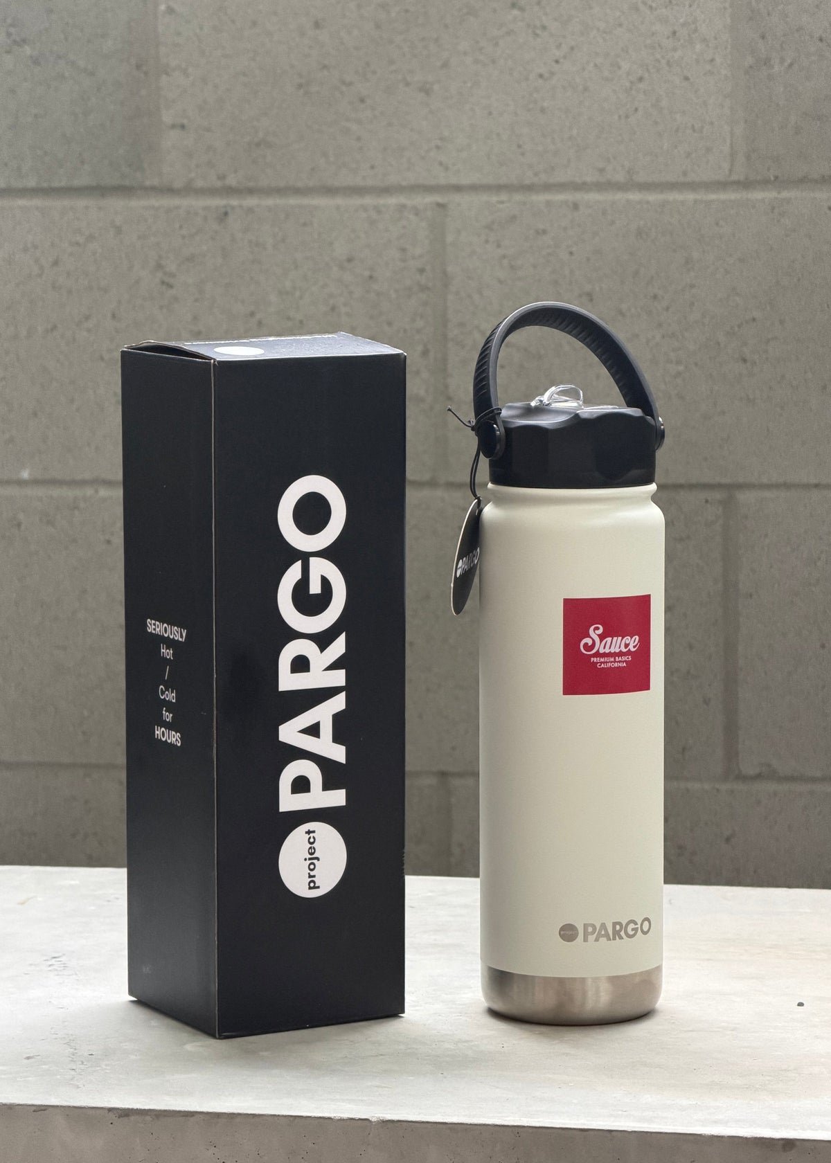 A cream-colored insulated water bottle with a black lid and carry loop stands next to its black Pargo packaging box on a light stone surface against a gray brick wall.