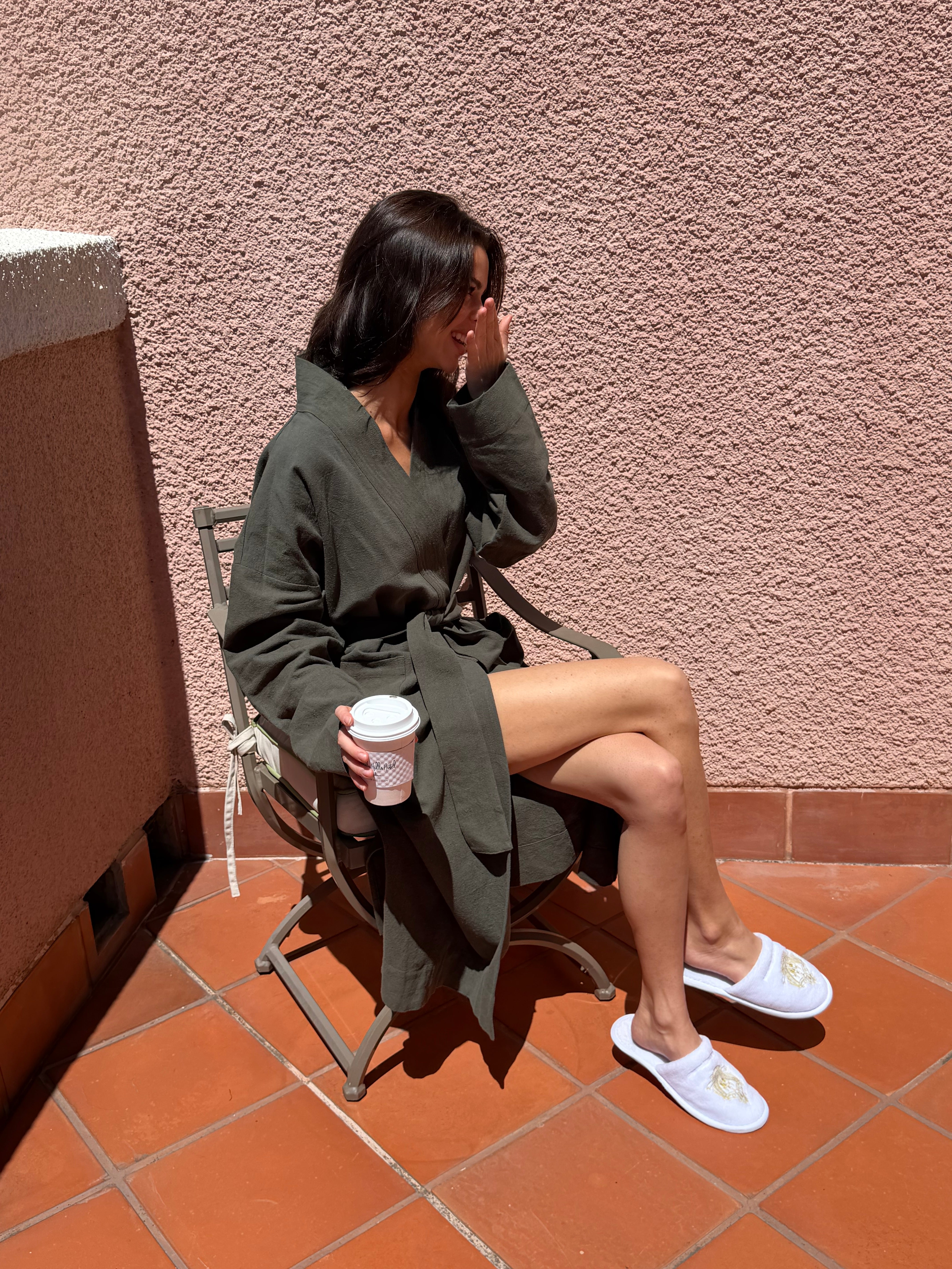 Woman in a robe sitting on a chair holding a coffee cup against a pink wall.