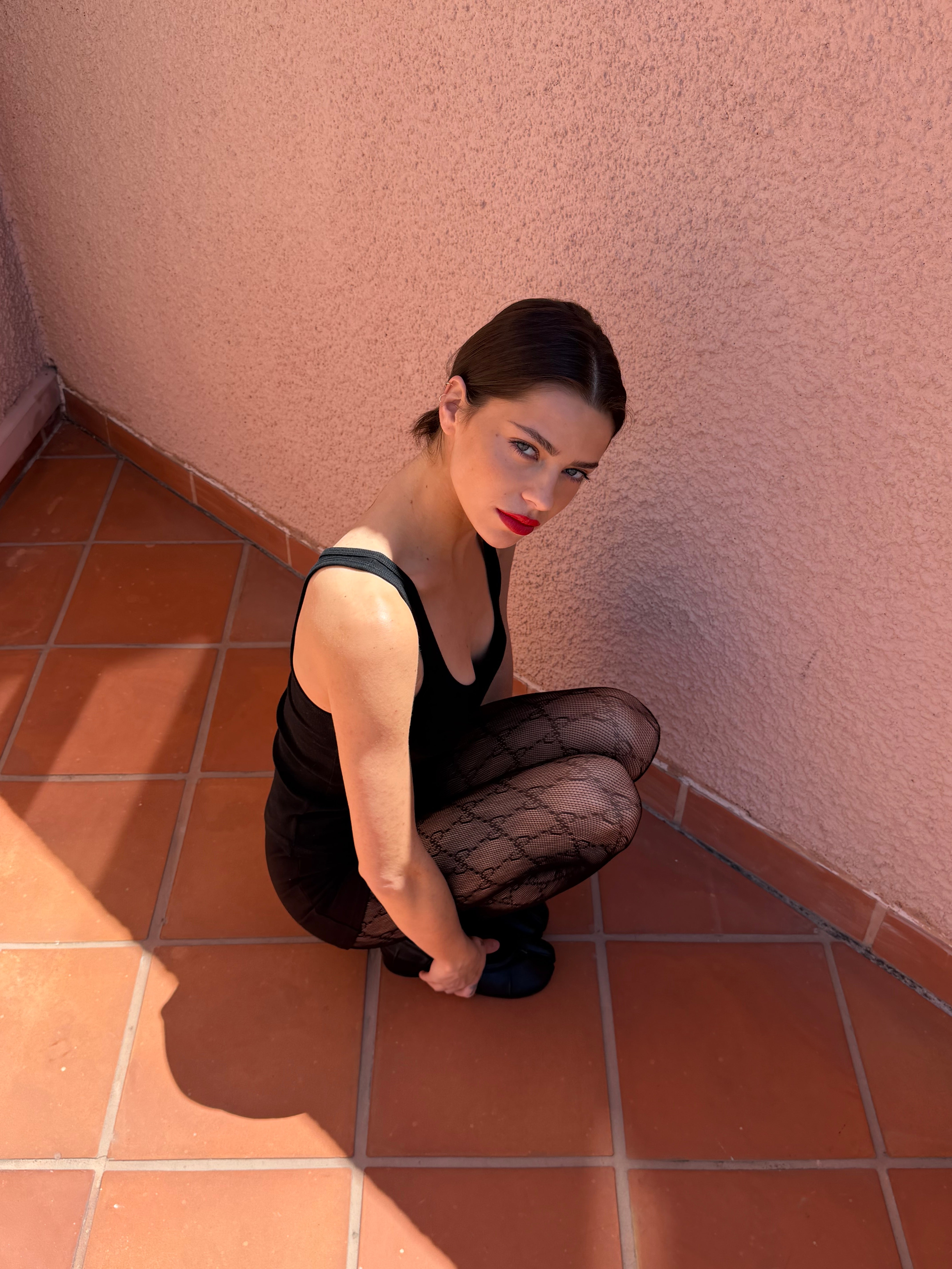 Woman in black outfit and fishnet stockings sitting on a tiled floor against a pink wall.