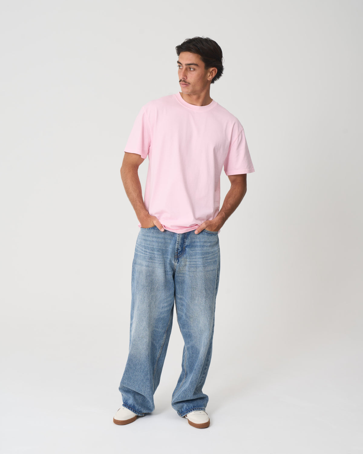 A man modeling a light pink crew-neck t-shirt and wide-leg blue jeans against a plain light gray studio background.