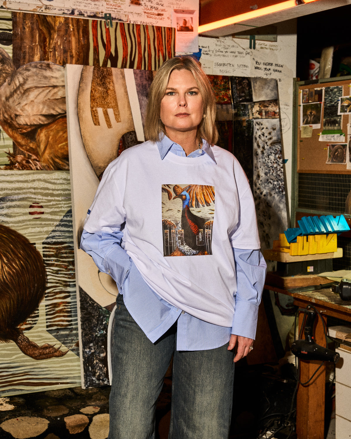 A woman stands in a creative studio wearing a light blue button-down shirt layered under a white oversized sweatshirt that features a colorful peacock graphic print on the front, paired with dark gray jeans.