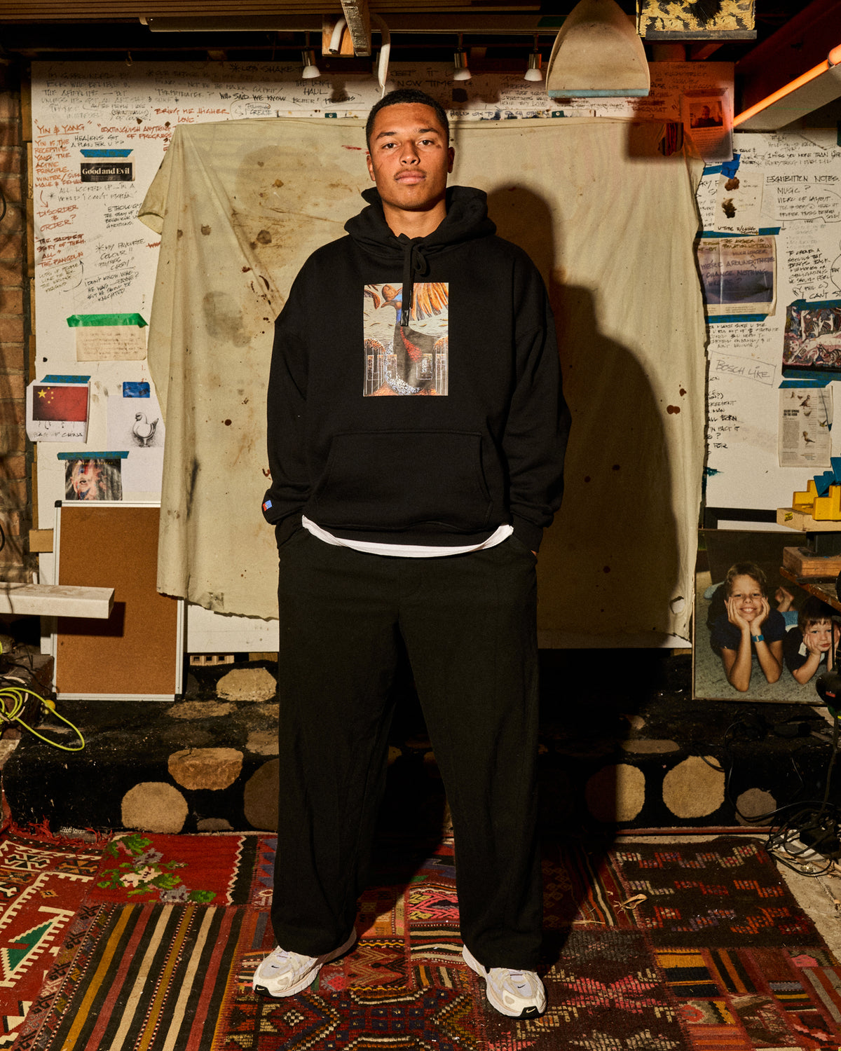 A man wearing a black pullover hoodie with a square graphic print on the chest, black sweatpants, and white sneakers, standing in a cluttered room with posters and a tapestry behind him.