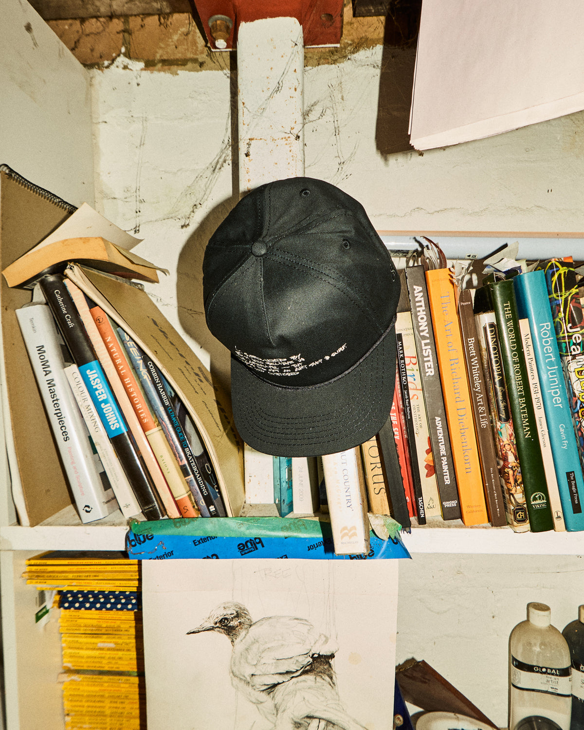 A black baseball cap with white embroidered text sits on a bookshelf surrounded by books, papers, and other cluttered items.