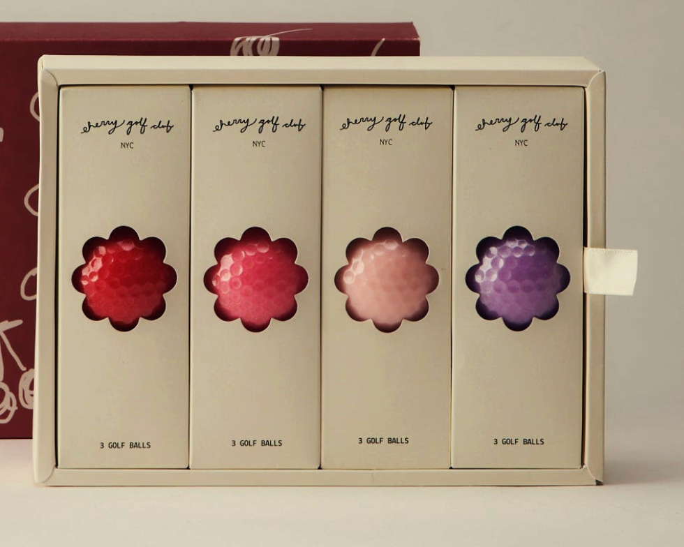 A boxed set of four golf balls in individual cream-colored packages, each with a flower-shaped cutout showing the golf ball color underneath. The visible ball colors are red, pink, peach, and purple.