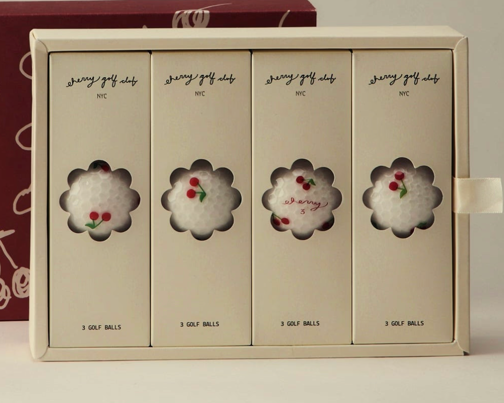 A boxed set of four golf balls in elegant cream-colored packaging, each panel featuring a flower-shaped cutout revealing a white golf ball with red cherry embellishments. The box is branded 'sassy golf clubs' with 'NYC' and '3 GOLF BALLS' printed on the panels.