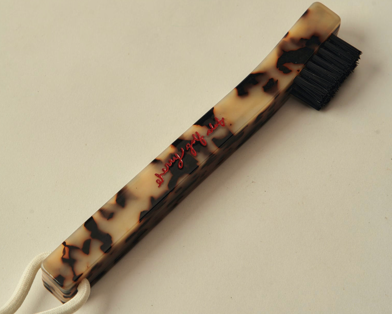 A vintage-style cleaning brush with a tortoiseshell-patterned handle, black bristles, and a white hanging loop, photographed on a plain light background.