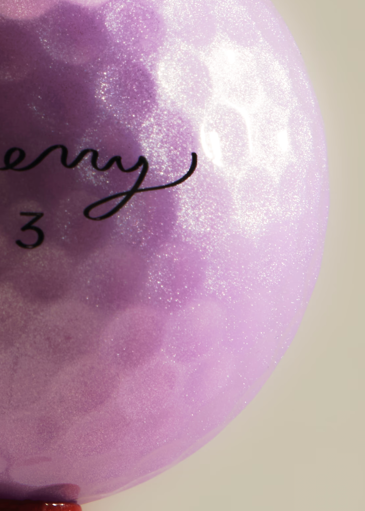 Close-up of a lavender-purple golf ball with a textured, hammered finish and black custom engraving, set against a solid light beige background.