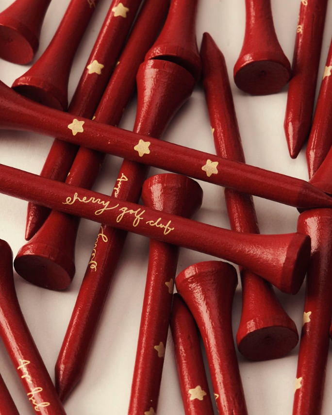 A pile of red wooden golf tees with gold star and script lettering, arranged on a white background.