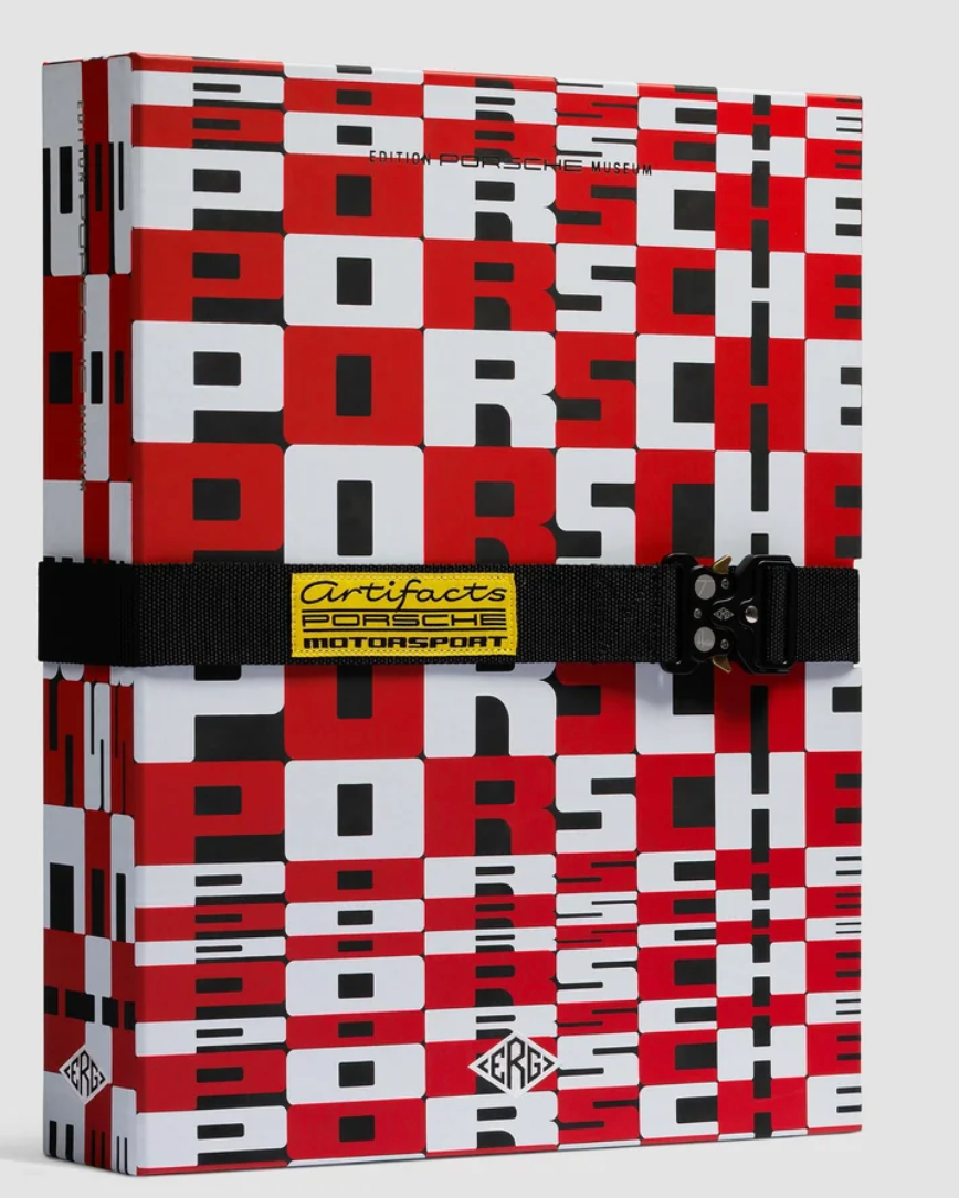 A Porsche-branded hardcover notebook or journal featuring a bold red, white, and black checkerboard pattern. It is secured with a black elastic band across the middle, which has a yellow and black Certificate of Authenticity label.