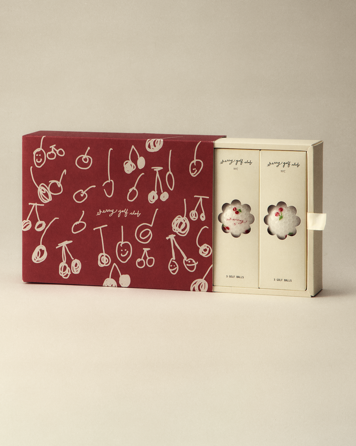 A boxed set of two small round pastries in cream-colored packaging, shown next to a larger dark red outer box decorated with white cherry illustrations, on a plain warm beige studio background.