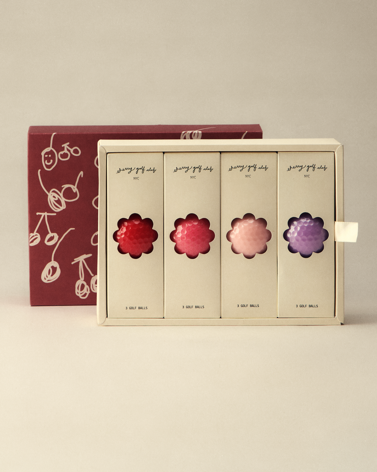 A boxed set of four flower-shaped cosmetic or fragrance items displayed in a cream-colored tray, with a dark red patterned box behind it on a light beige studio background.