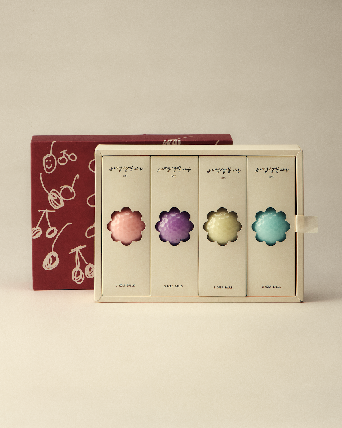A boxed set of four flower-shaped cosmetic or candy pieces in pastel pink, purple, yellow, and blue, displayed in front of a dark red box with white cherry illustrations on a warm beige background.