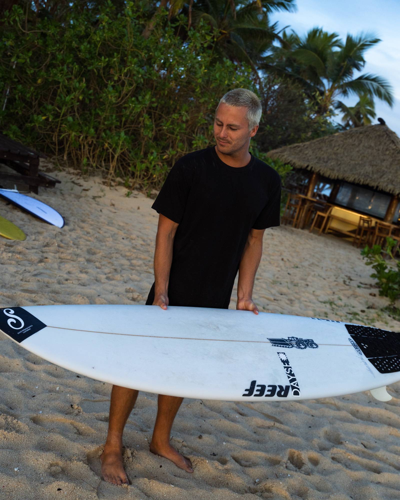 Man holding a surfboard on a beach with palm trees and a hut in the background