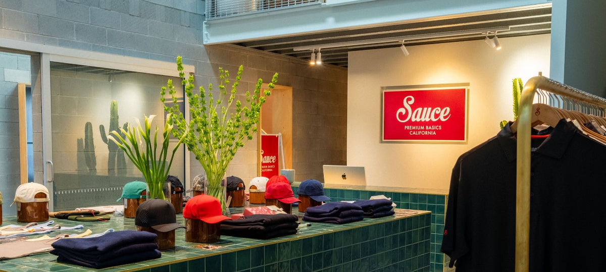 A retail clothing display featuring a green tiled counter with folded shirts, hats, and a plant, alongside a clothing rack with a black shirt. A red sign on the wall reads 'Sauce' with 'Mission-Based Outfitter' below it.
