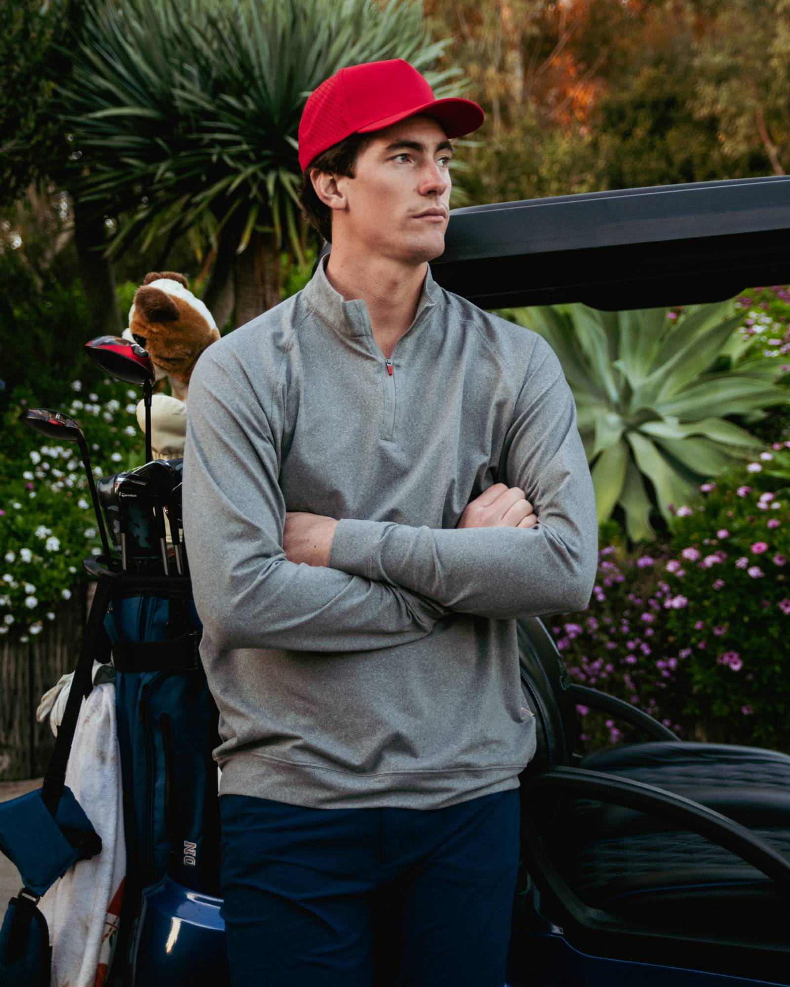 Man in gray jacket and red cap standing next to a golf bag with golf clubs, outdoors.