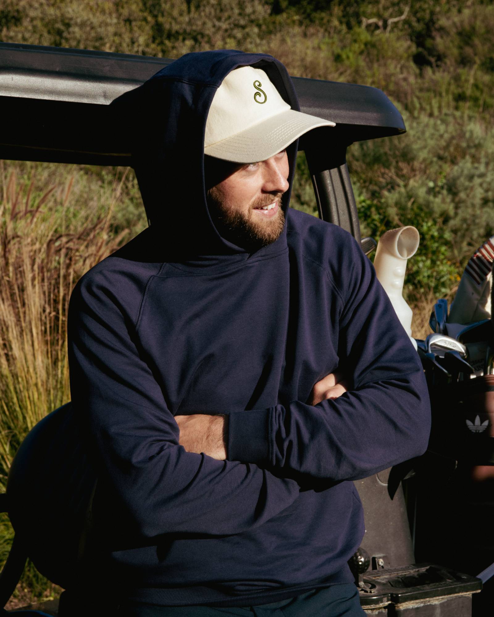 Man wearing a navy blue hoodie and white cap standing next to a vehicle outdoors.