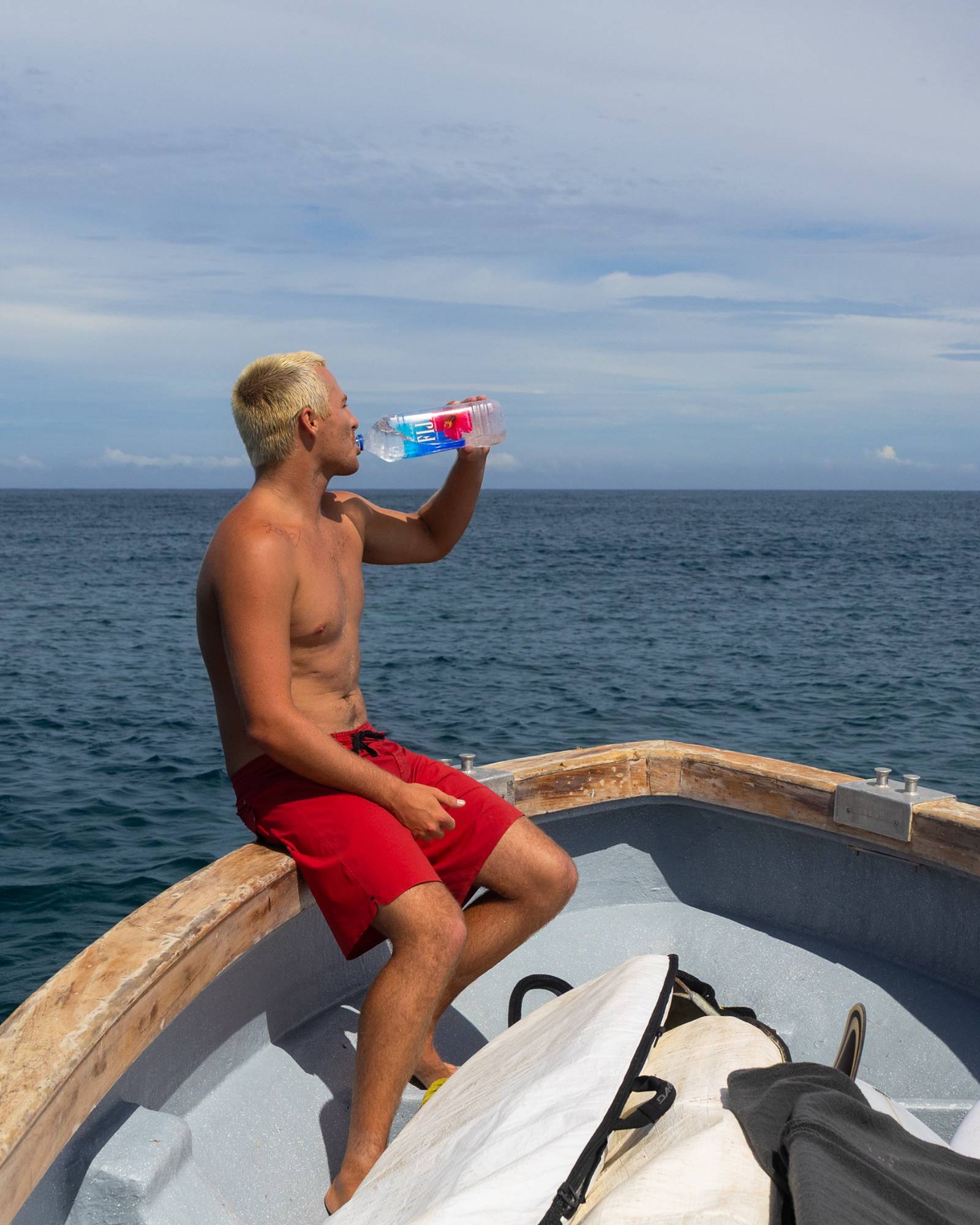 Man in red shorts sitting on a boat, drinking from a bottle with a blue sky and ocean background.