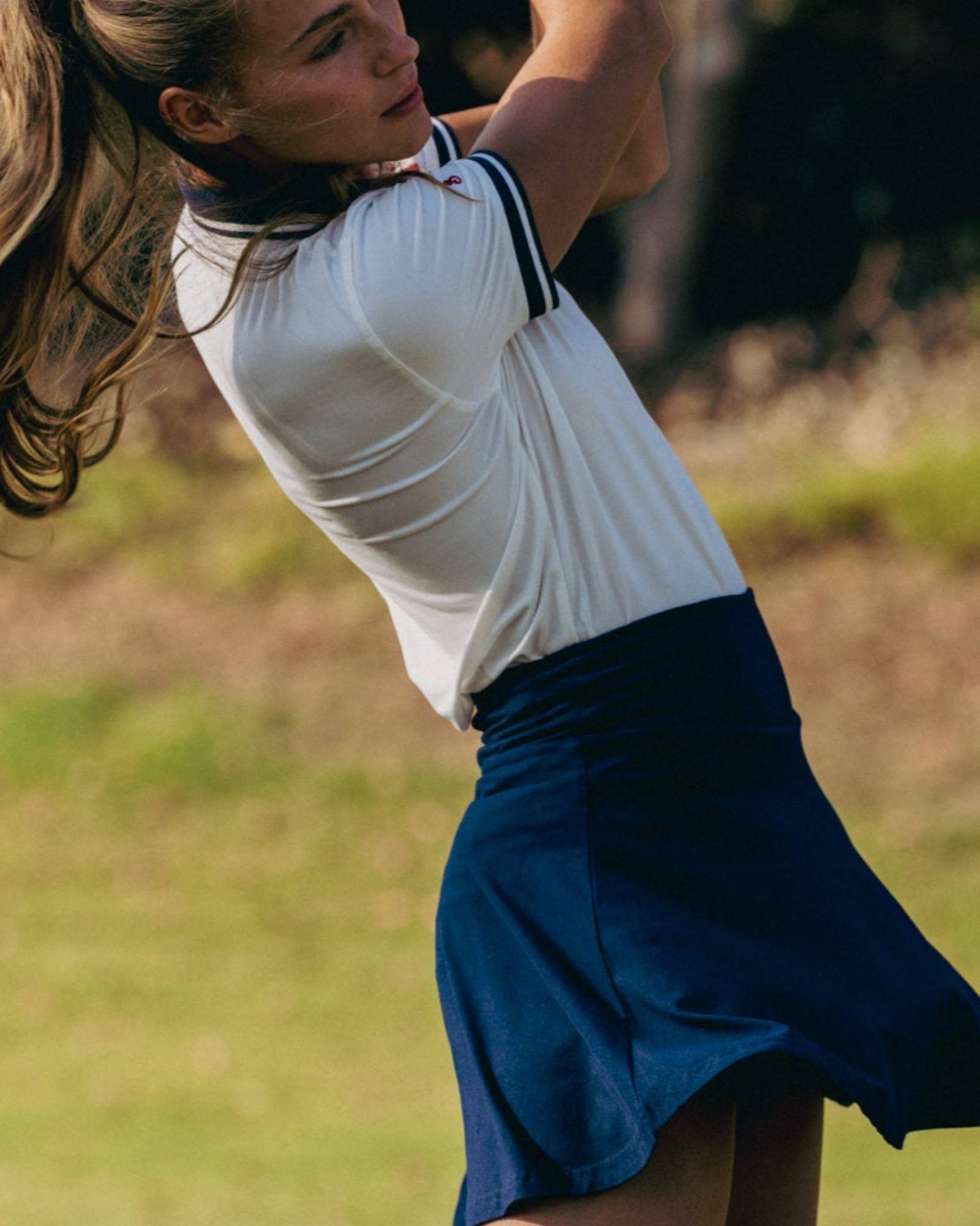 Woman in a white top and blue skirt swinging a golf club on a golf course.