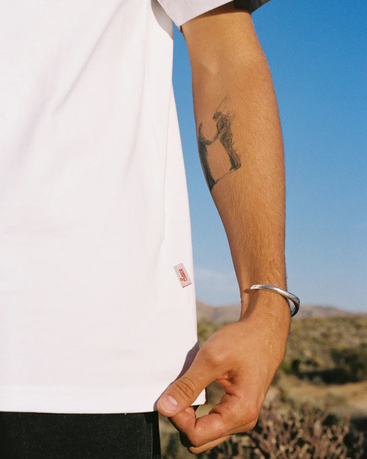 Person wearing a white t-shirt with 'Sauce' logo, against a scenic background.