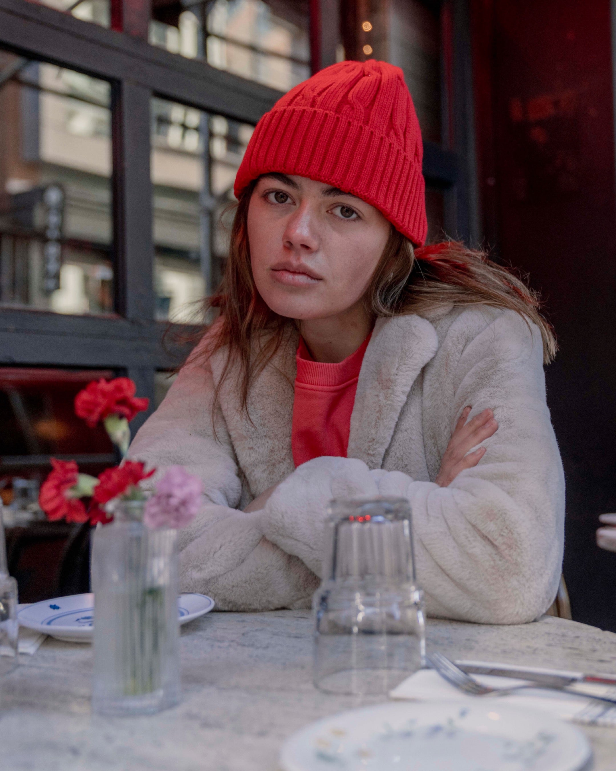 Person wearing a red beanie and light-colored coat sitting at a table with a vase of flowers and glassware.