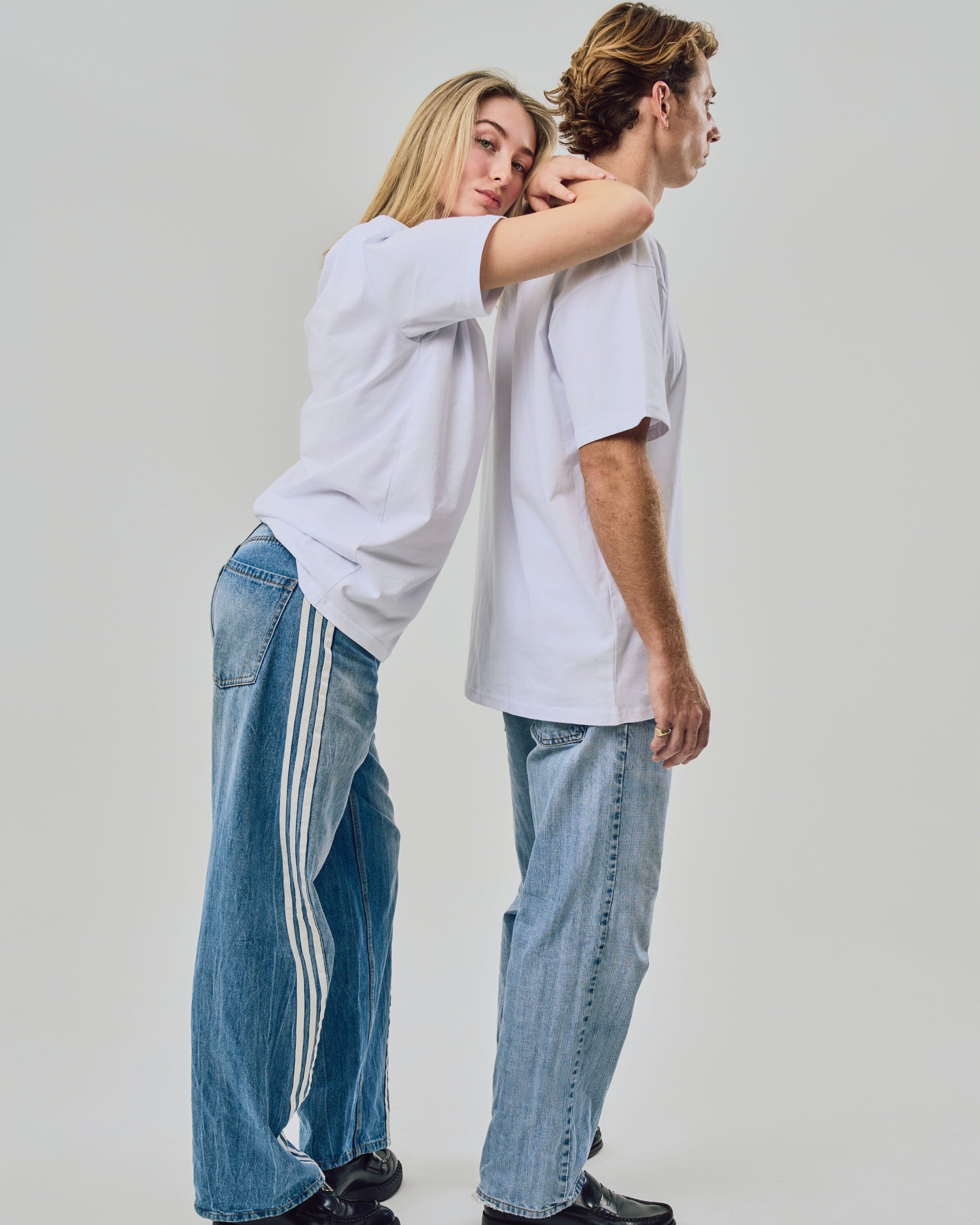 Two people wearing matching white t-shirts and blue jeans with white stripes on a plain background