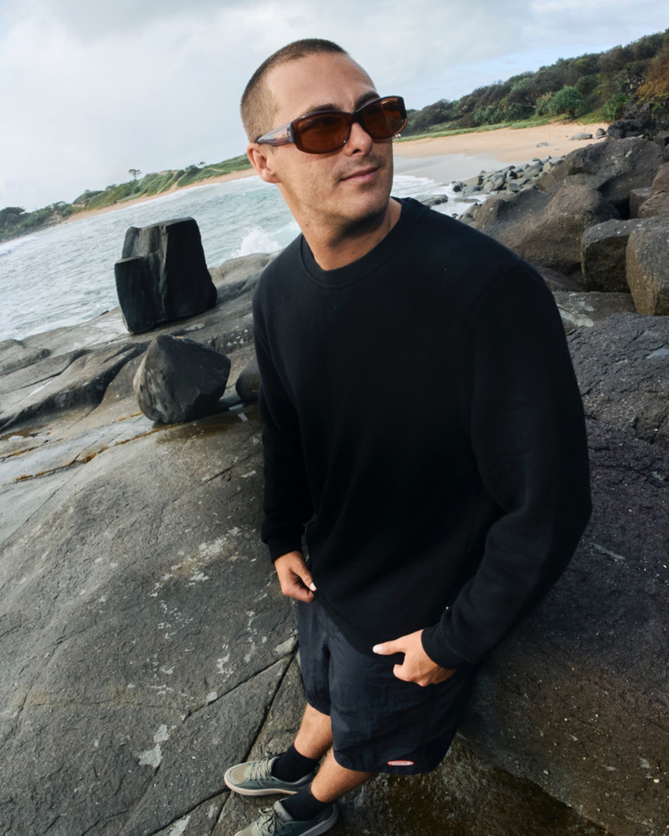 Man wearing sunglasses and a black outfit standing on a rocky beach with water and trees in the background.