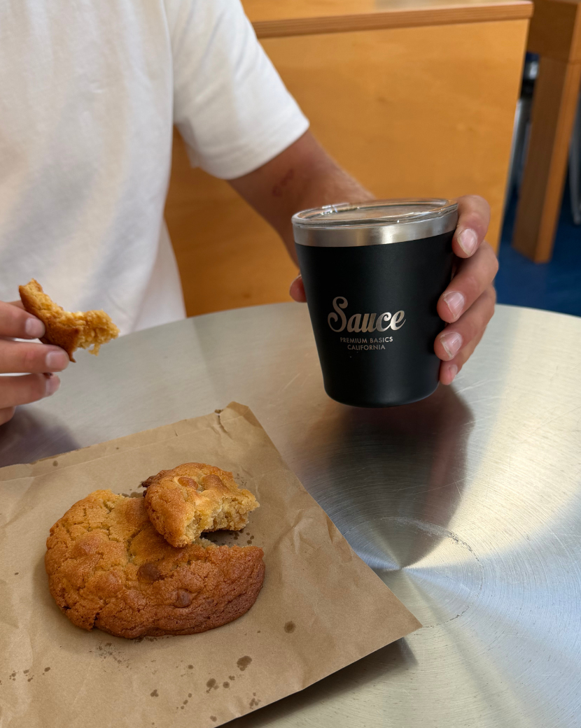 A person in a white shirt holds a black insulated tumbler with the word 'Sauce' printed on it, while a half-eaten pastry sits on brown paper on a stainless steel table.