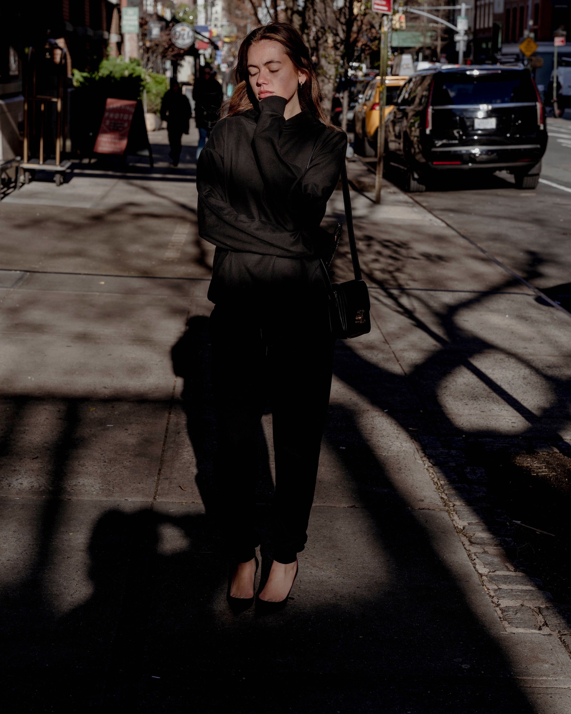 Woman in black outfit walking on a city street with shadows cast by trees.