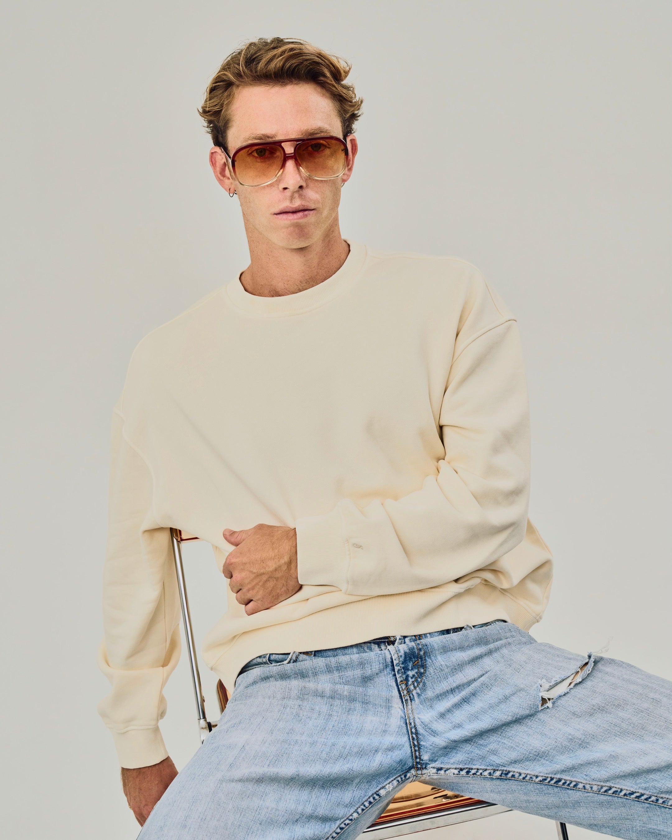 Man wearing a beige sweater and light blue jeans sitting on a chair against a plain background