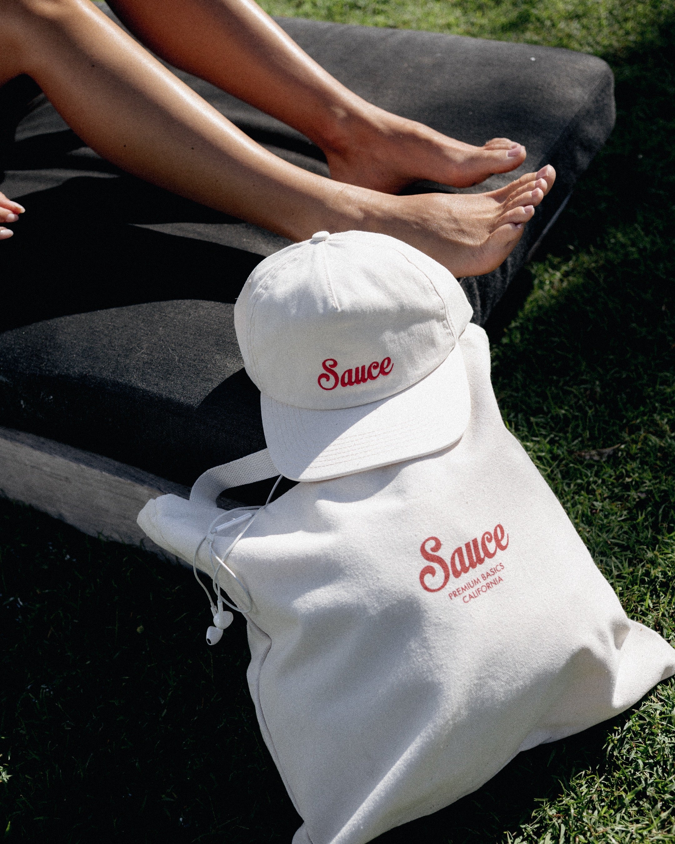 White tote bag and cap with 'Sauce' logo on grass