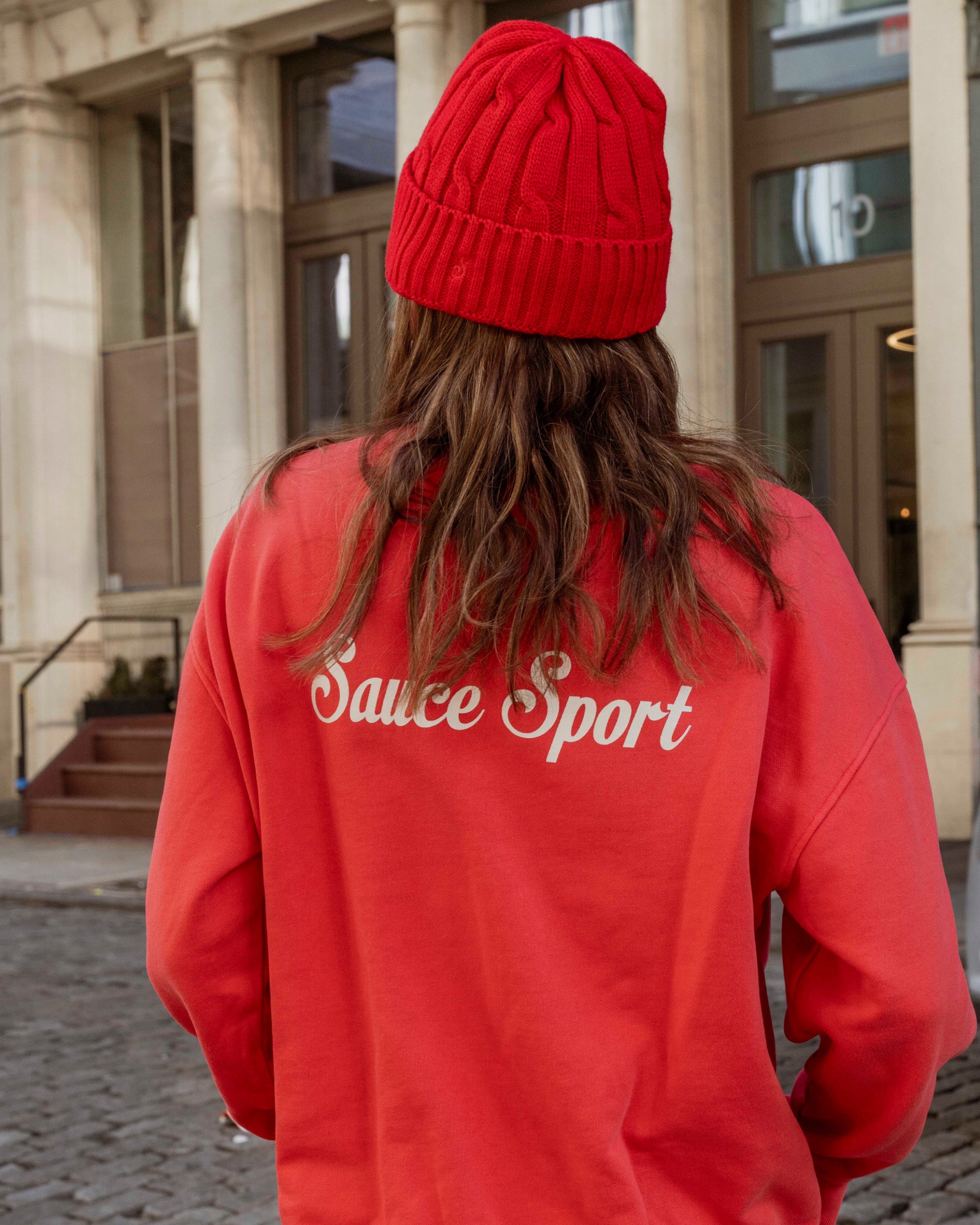 Person wearing a red 'Sauce Sport' sweatshirt and beanie outdoors.