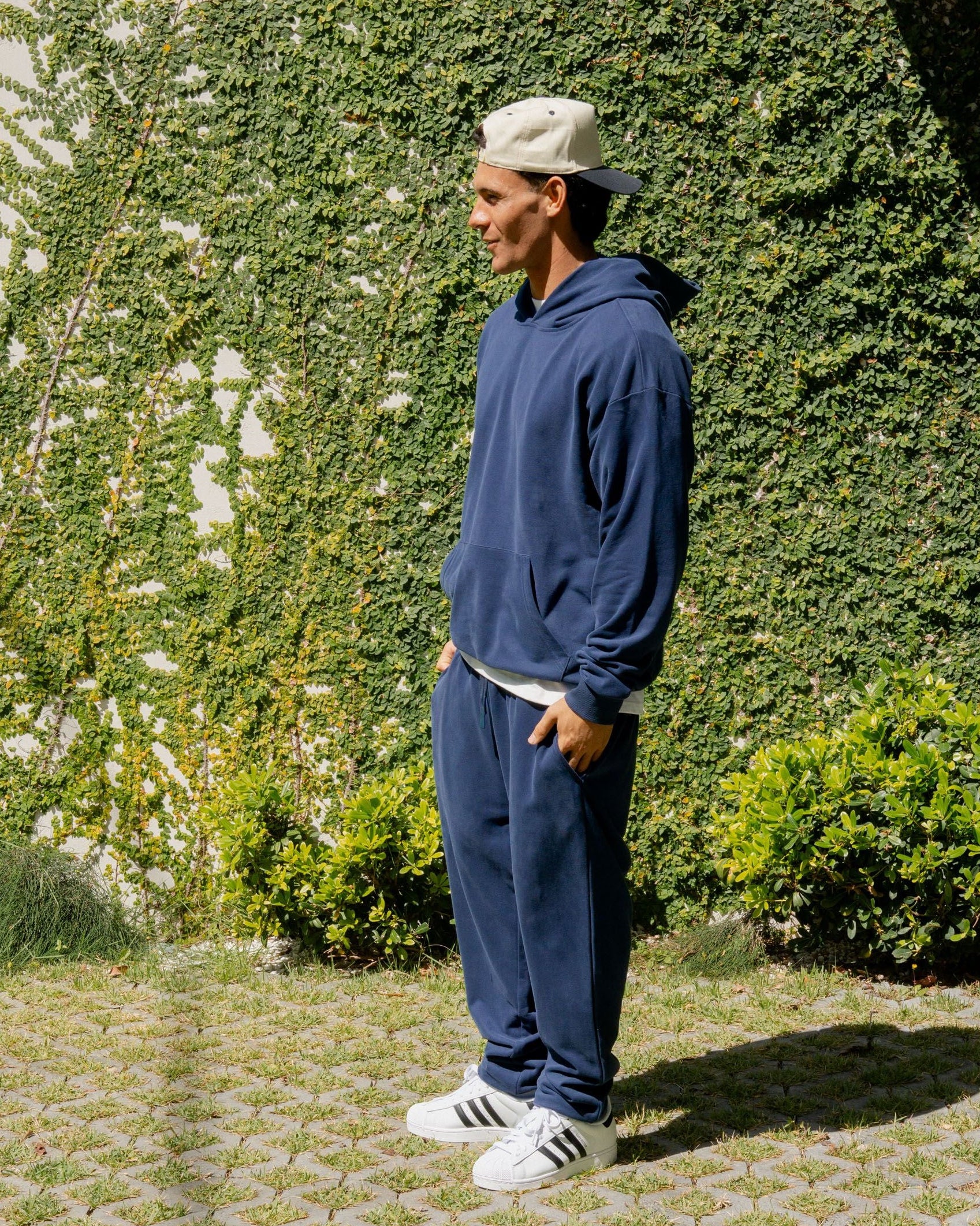 Person wearing a blue tracksuit and white sneakers standing in front of a green hedge.
