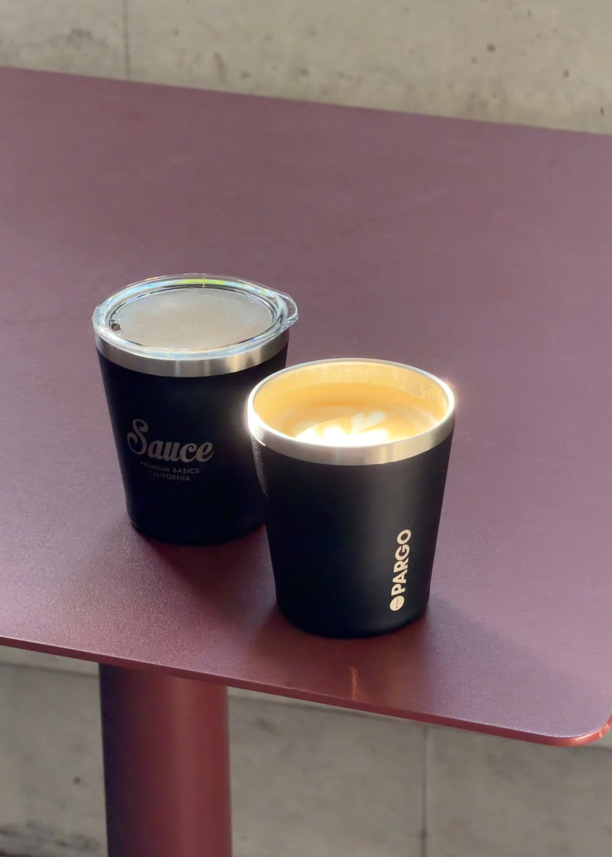 Two black insulated travel cups on a reddish-brown table, one with a clear lid and the other open with a flame, both branded Pargo. The background shows a concrete wall.