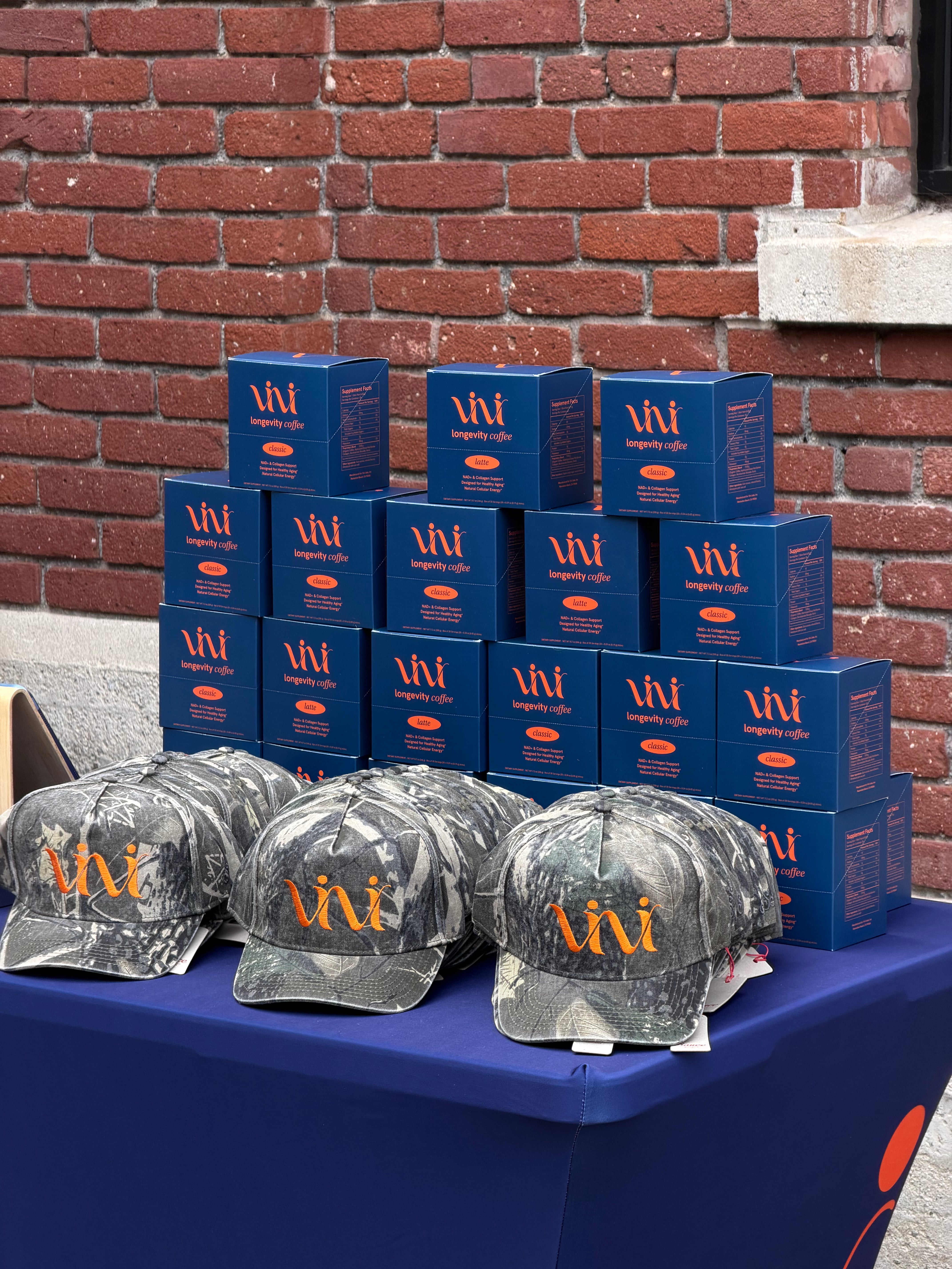 Stacks of blue boxes with a logo and camo hats on a table against a brick wall.