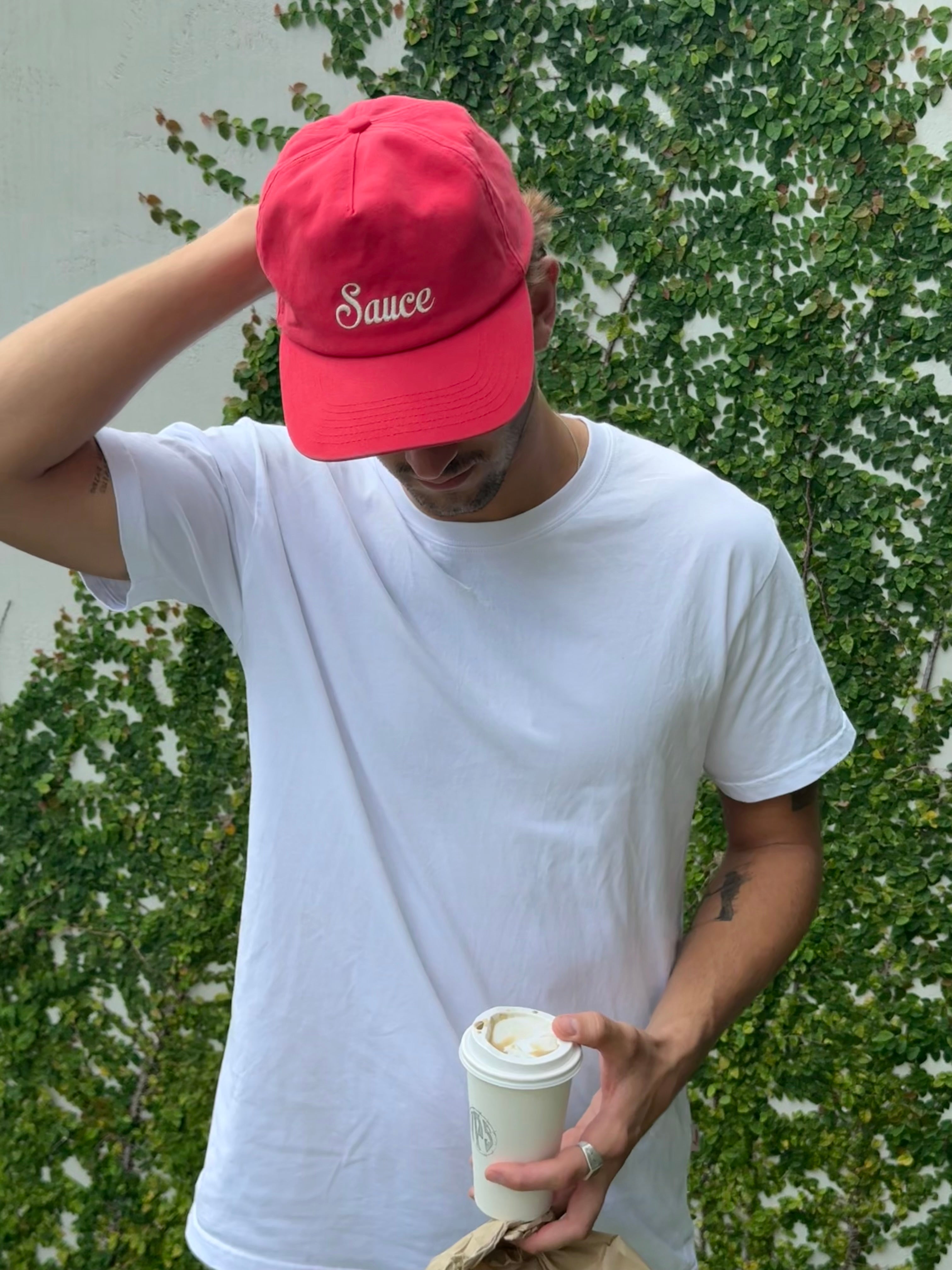 Person wearing a red cap with 'Sauce' text, holding a coffee cup against a green leafy background