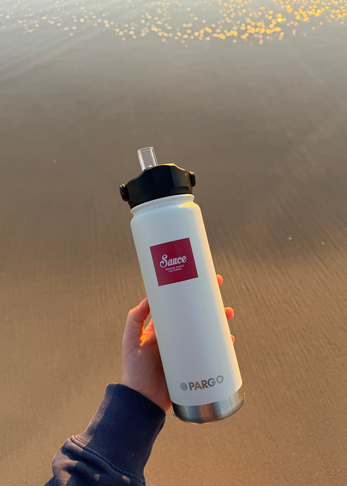 A hand holding a white Pargo insulated water bottle with a black lid and clear straw, set against a blurred beach background at sunset.