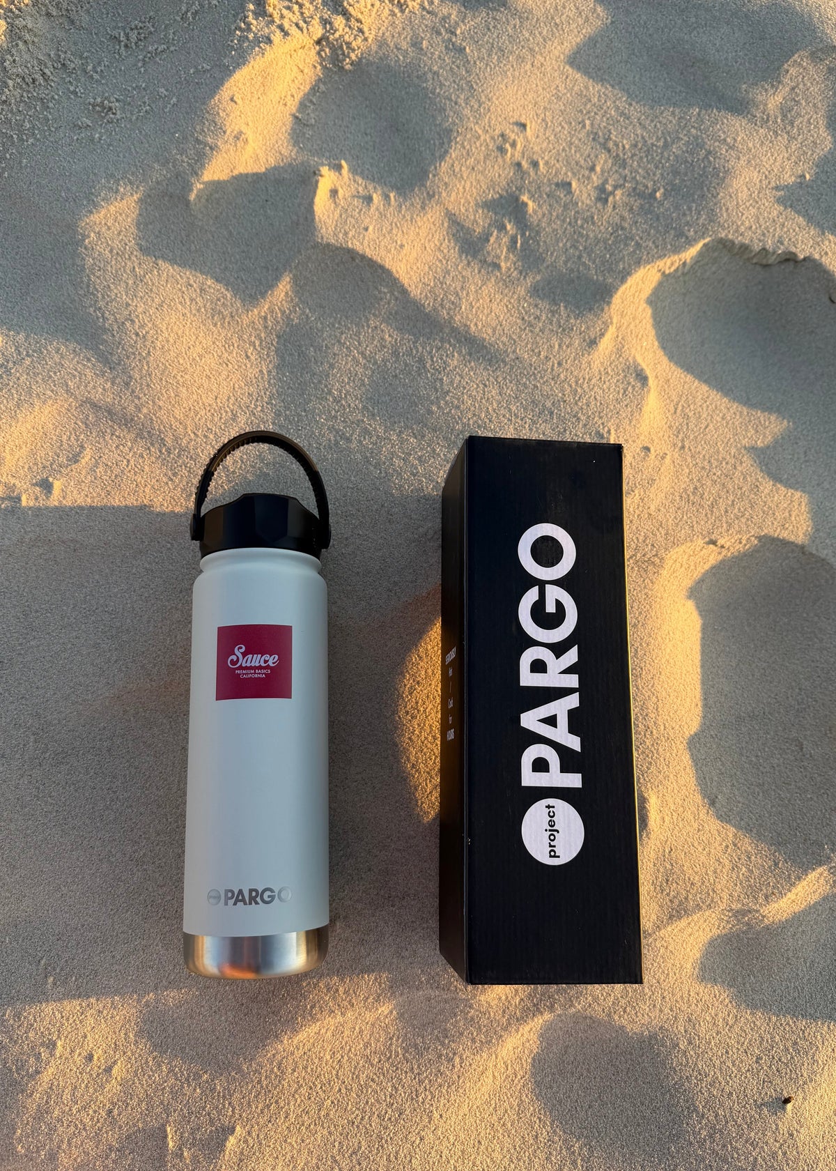 A white insulated water bottle with a black loop cap and a small red logo label sits beside a tall black PARGO box on sandy ground in warm golden-hour light.