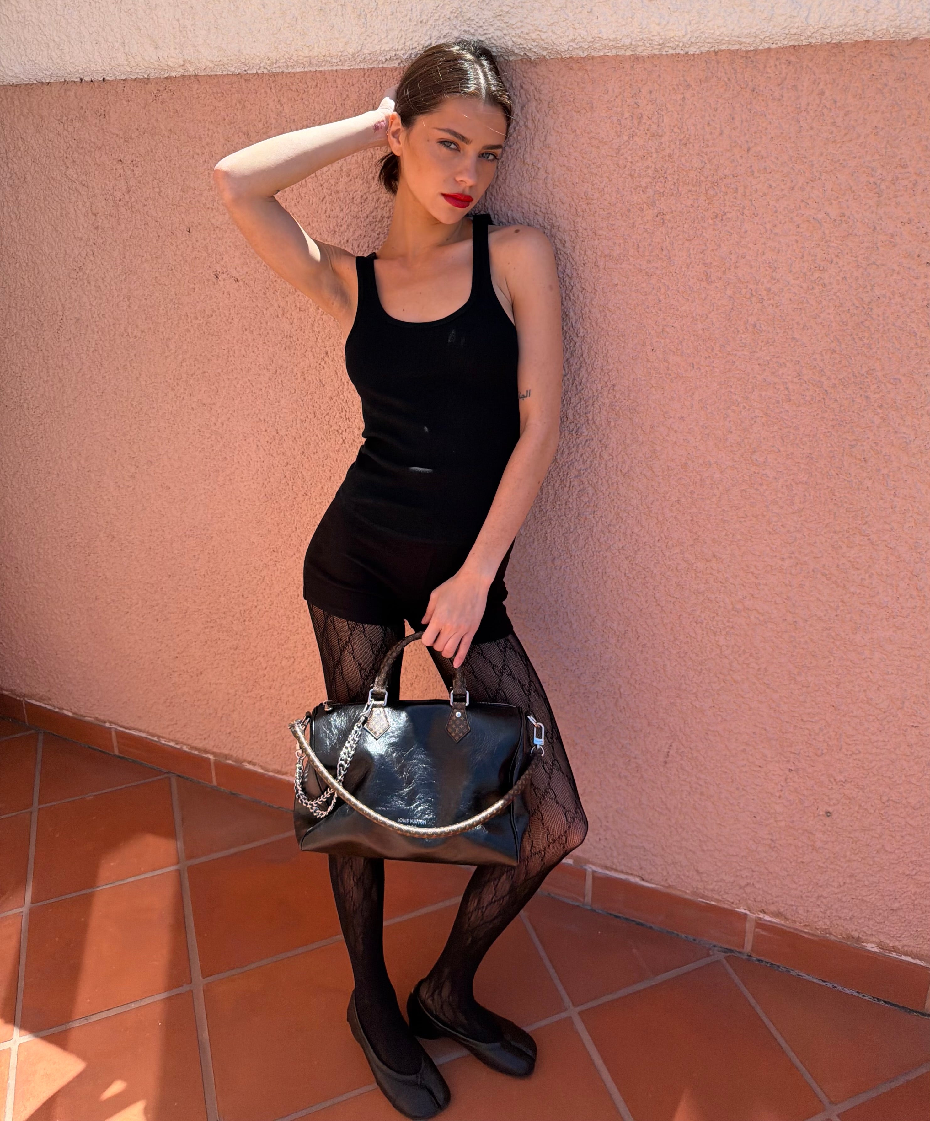 Woman in a black tank top and shorts  holding a handbag against a pink wall.