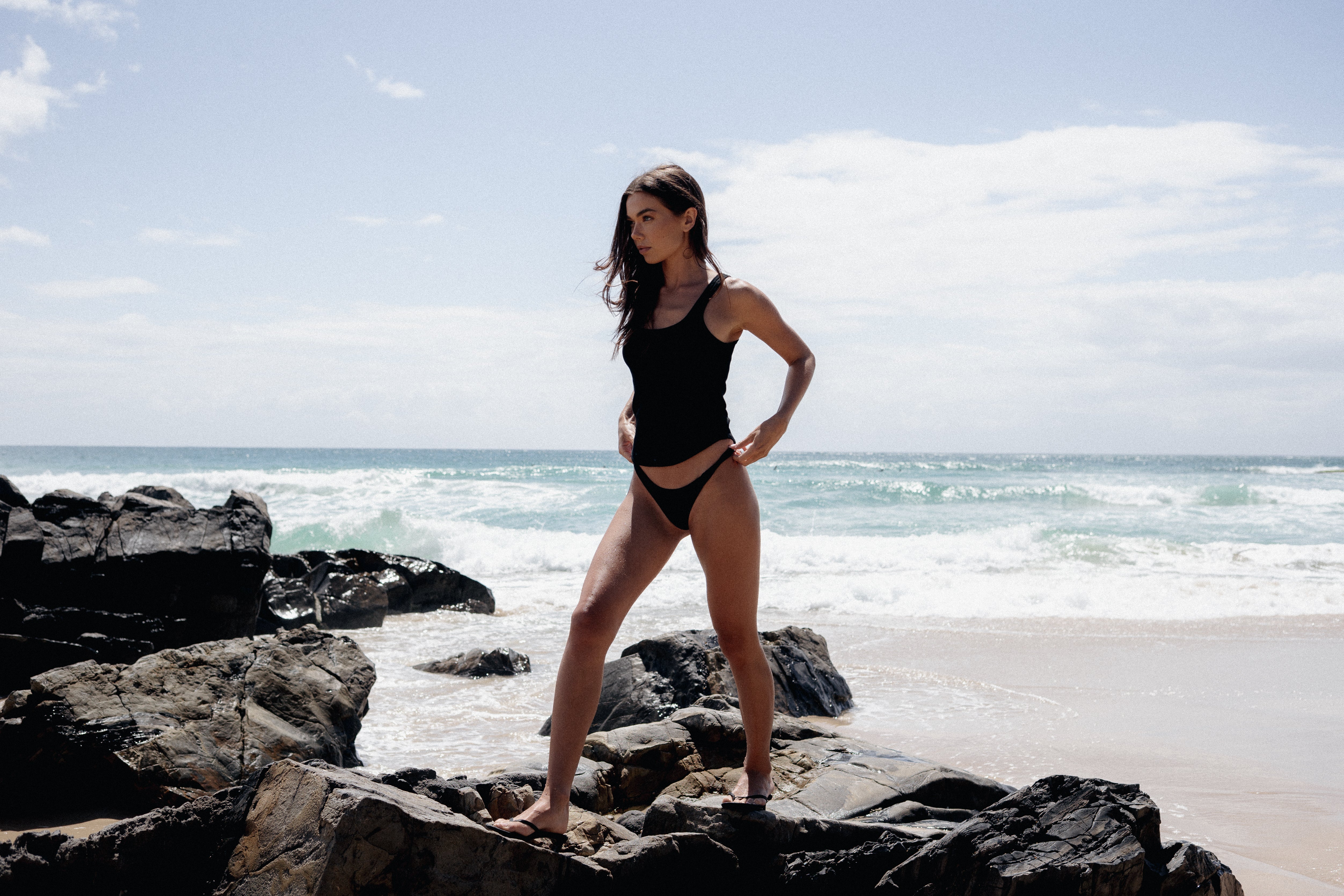 Woman in a black tank top standing on rocks by the ocean