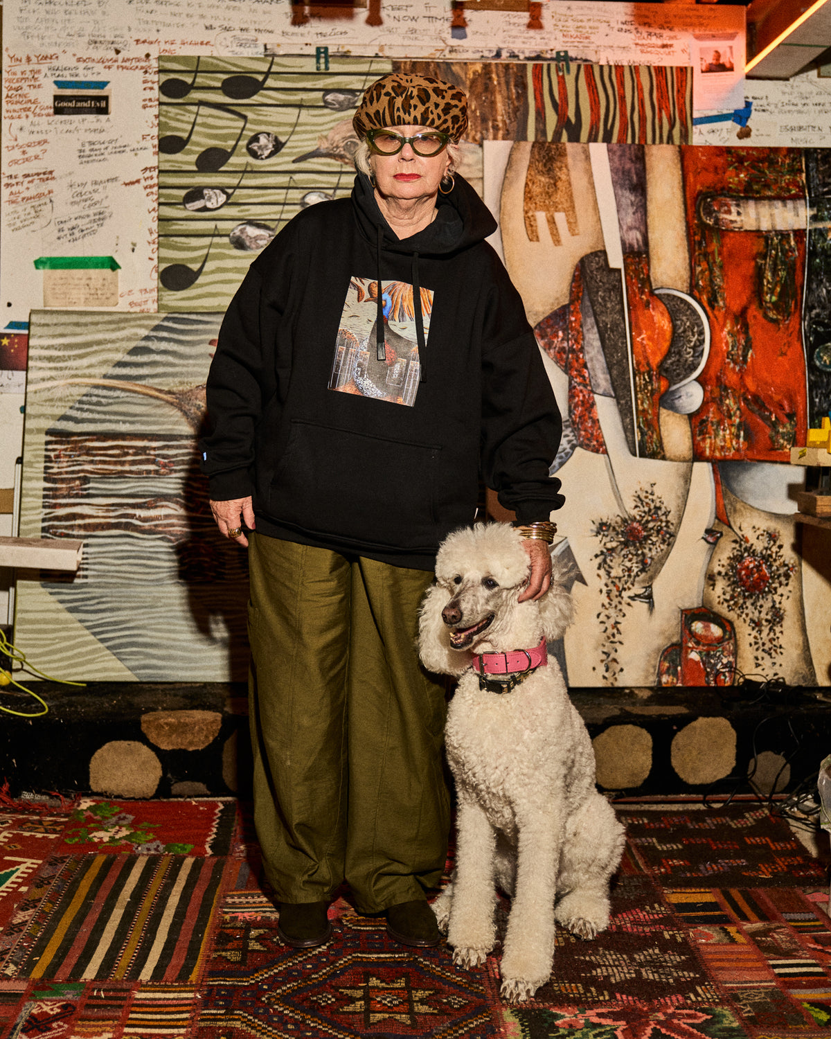 A person wearing a black graphic hoodie, olive green pants, and a leopard-print hat stands indoors next to a white poodle. The background is a busy art studio or gallery space with paintings, rugs, and papers.