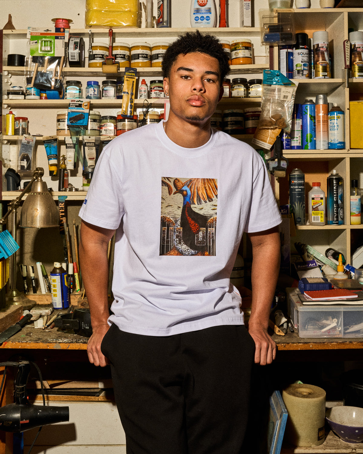 A man wearing a white t-shirt with a colorful graphic print of a peacock, standing in a cluttered art studio filled with shelves of paint cans and supplies.