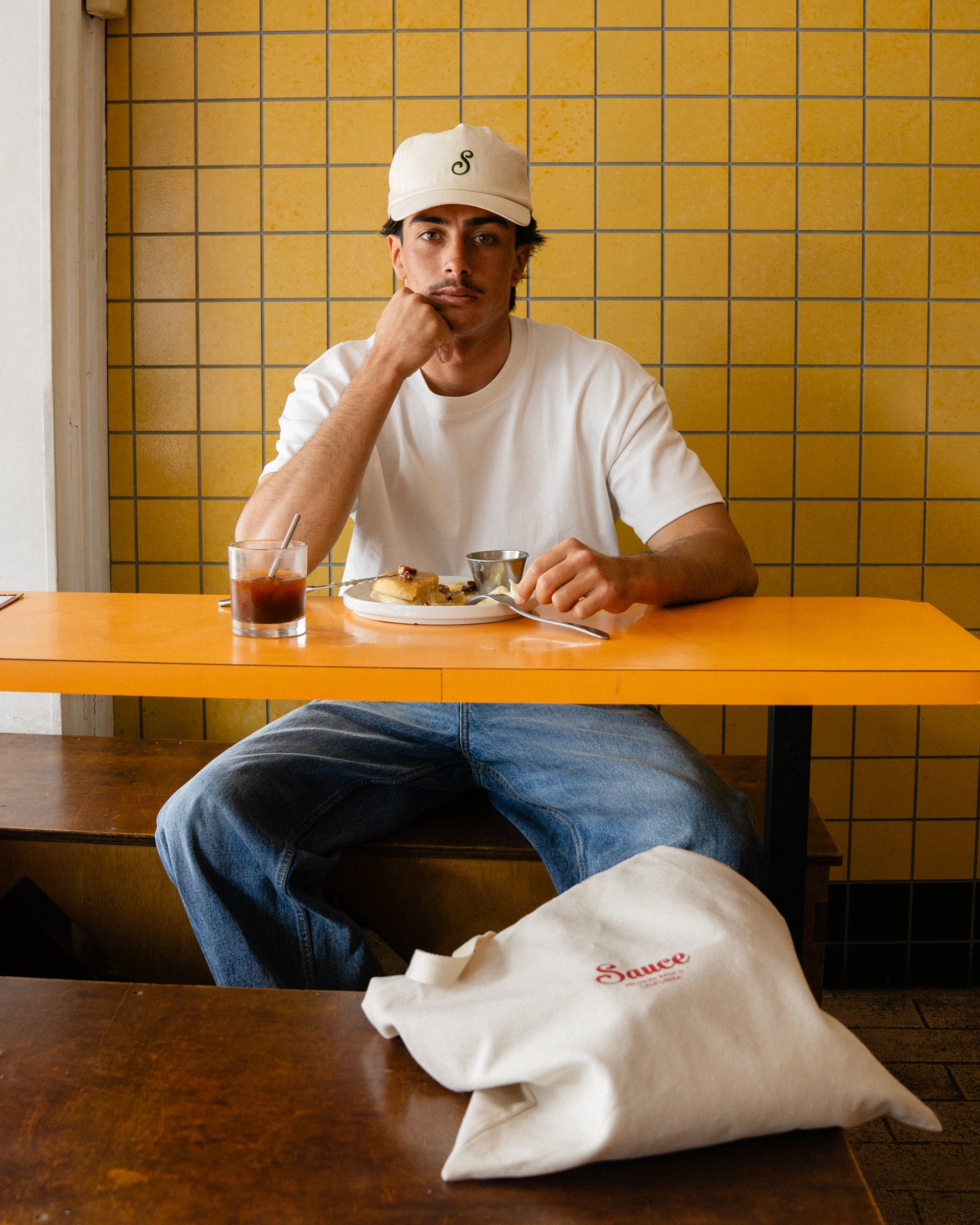 Man sitting at a table with a plate of food and a drink, wearing a white t-shirt and cap, against a yellow tiled wall.