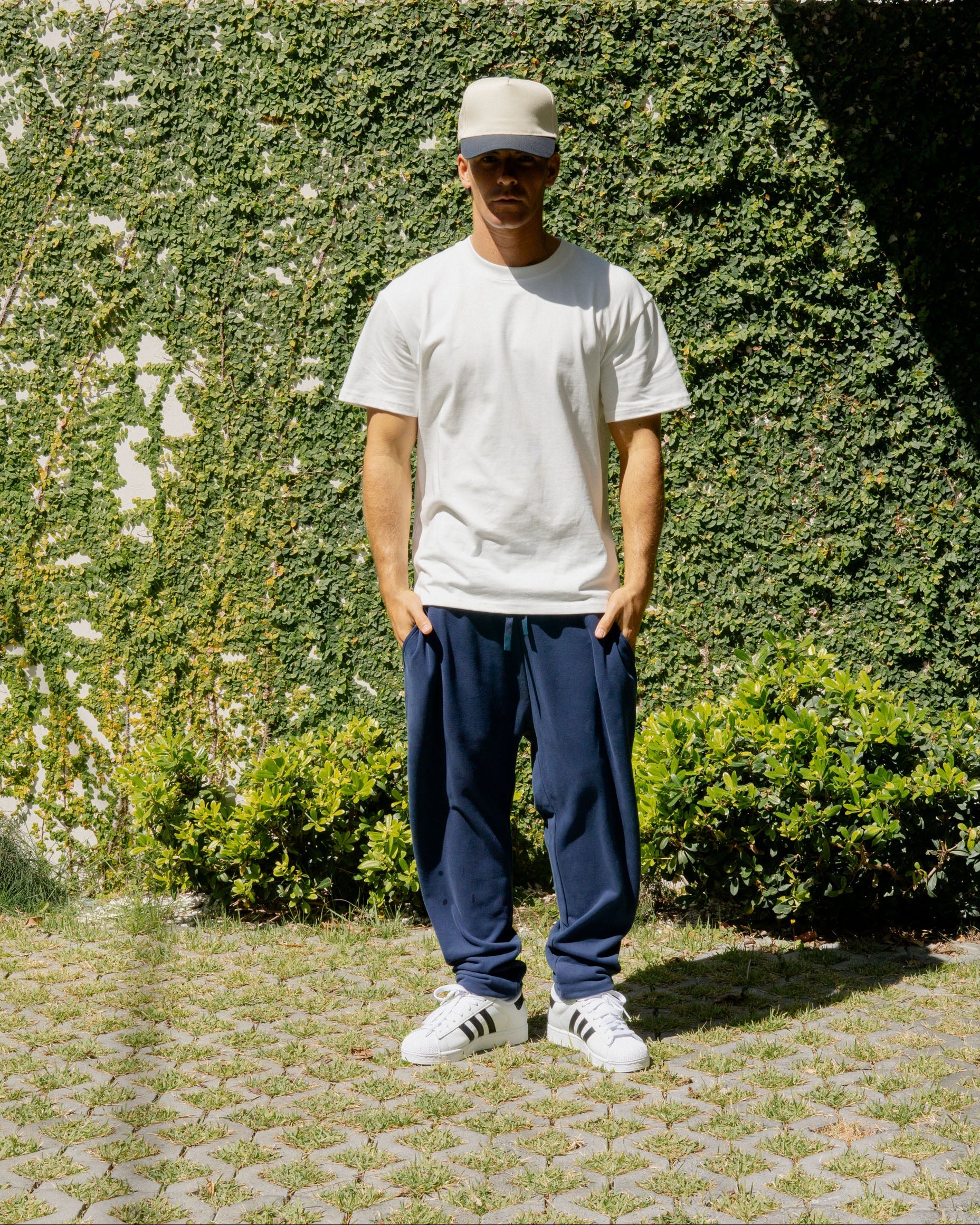 Person wearing a white t-shirt, navy pants, and white sneakers standing in front of a green hedge.