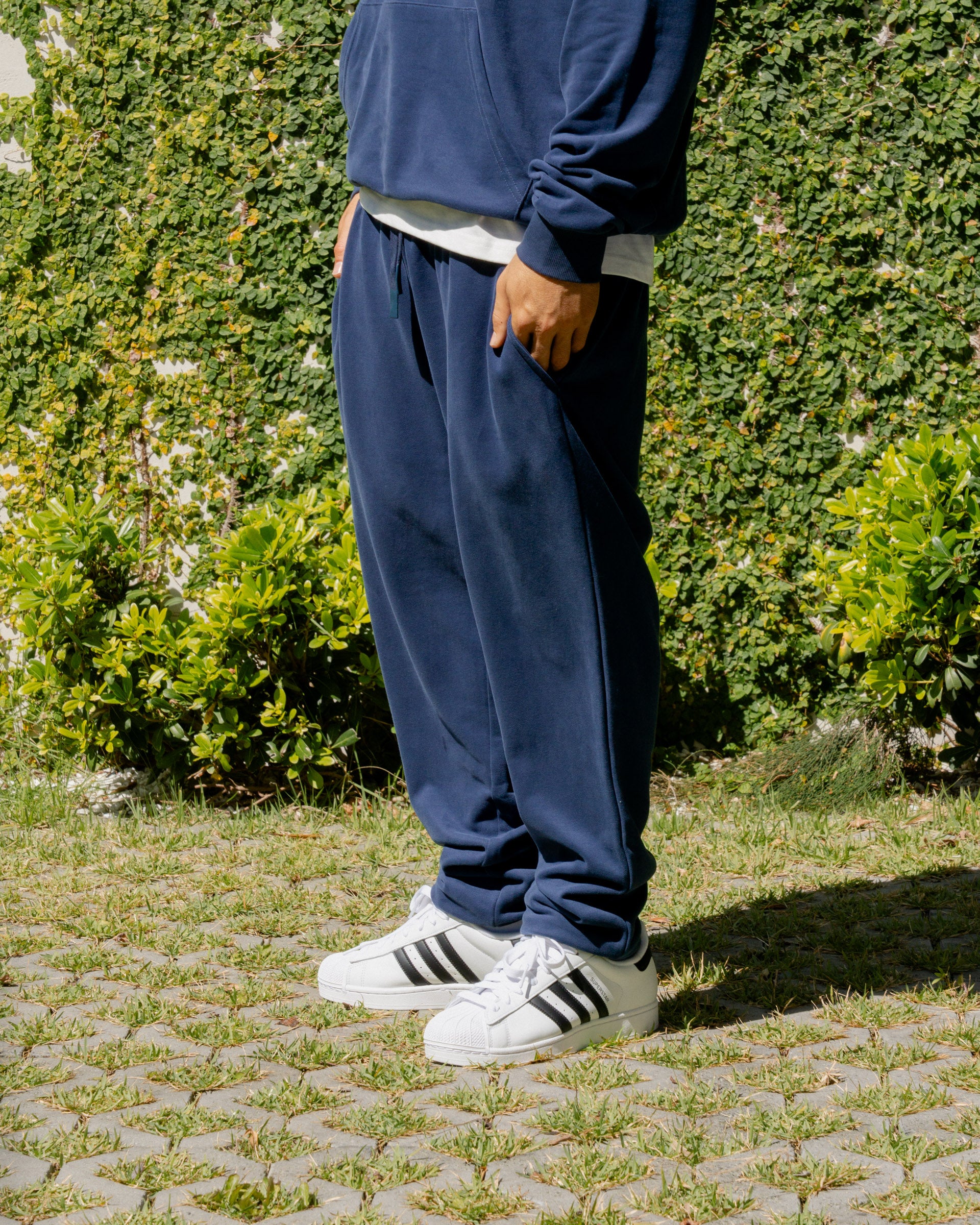 Person wearing navy blue sweatpants and white Adidas sneakers standing on a stone path with greenery in the background.
