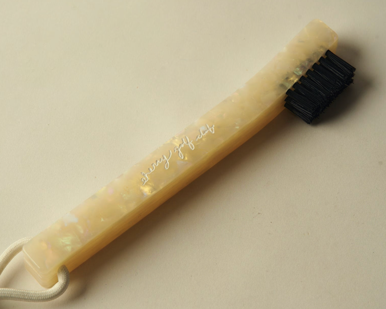 A small cleaning brush with a translucent iridescent cream handle and black bristles, photographed on a plain beige background. The handle has a script logo printed on it and a white loop at one end.